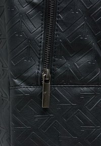Sac en cuir noir texturé avec une fermeture éclair proéminente et un tirette en métal, arborant un motif géométrique embossé sur la surface.