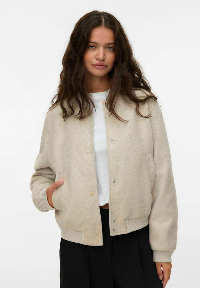 Giacca bomber beige realizzata in tessuto morbido, con chiusura a bottoni a pressione, polsini a coste e tasche laterali, indossata sopra una camicia bianca.
