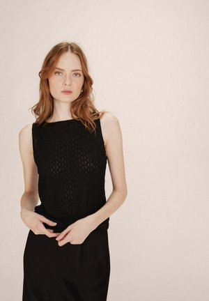 Jeune femme aux cheveux roux portant un haut sans manches noir à motifs et une jupe noire devant un fond beige uni.