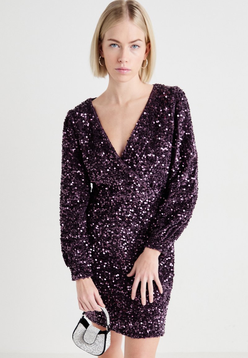 Fioletowa mini sukienka z cekinami, z długimi rękawami i głębokim dekoltem w kształcie litery V. Materiał ma błyszczącą fakturę. W komplecie z małą srebrną torebką.