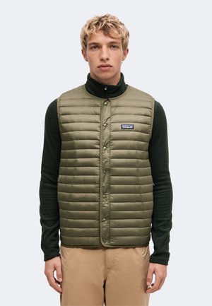 Jonge man met kort krullend blond haar, gekleed in een olijfgroene doorgestikte bodywarmer over een donkergroen shirt met lange mouwen en een beige broek, recht voor zich uitstaand.