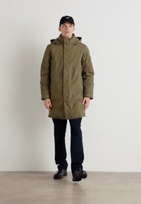 Olivgrüner gepolsterter Parka mit Kapuze, Reißverschluss vorne und Seitentaschen, kombiniert mit dunklen Hosen und schwarzen Schuhen vor einem schlichten Hintergrund.