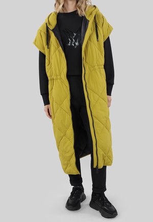 Persoon draagt een lange, mouwloze gele gewatteerde capuchonvest over een zwarte trui, broek en zwarte chunky sneakers.