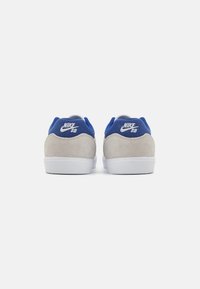 Nike SB apavi ar pelēku zamšādu augšdaļu, zilu apkakli un baltu gumijas zoli. "Nike SB" logo redzams uz papēža.