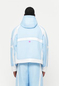 Niebieska i biała kurtka z kapturem o teksturowanym wykończeniu, z panelem siatkowym i różowymi akcentami. Przestronny krój ze luźnym dopasowaniem.