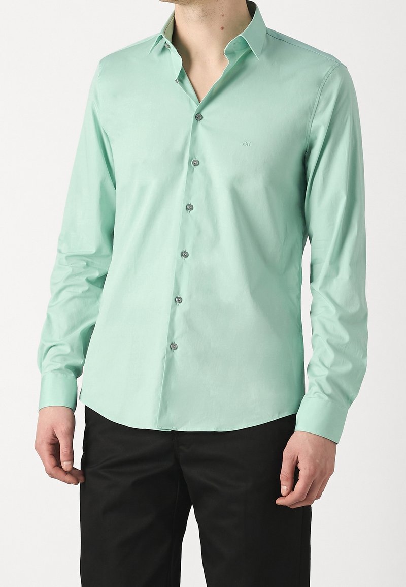 Homme portant une chemise à manches longues vert menthe avec un petit logo sur la poitrine et un pantalon noir, se tenant devant un fond uni.