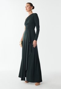 Donkergroene, lange mouwen, one-shoulder jurk gemaakt van gladde stof met een gedrapeerd ontwerp, met een gegolfde zoom en een clutch met gouden accenten.