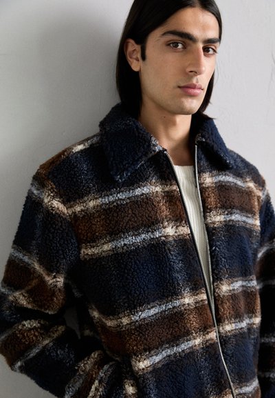Casaco de fleece xadrez castanho e azul-marinho com gola e fecho de zip, apresentando uma textura suave e riscas horizontais contrastantes.