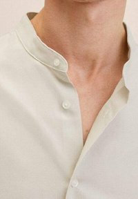 Camicia collare senza colletto di un beige chiaro, con piccoli bottoni bianchi e una texture di tessuto morbida. Design semplice e minimalista.