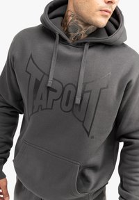Szürke kapucnis pulóver, amelyen elöl egy nagy fekete "TAPOUT" logó található. Zsinóros kapucnival és elülső kenguru zsebbel rendelkezik. Puha szövetből készült.