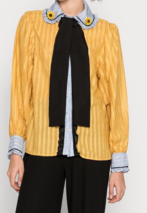 Chemisier rayé jaune avec des manches bouffantes, un col bleu à volants et un nœud papillon noir. Doté d'une fermeture à boutons à l'avant et de détails en dentelle noire.