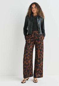 Czarna skórzana krótka kurtka z zapięciem na guziki, zestawiona z luźnymi, brązowymi spodniami w cętkowany wzór. Modelka nosi czarne sandały na paski.