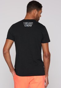 Svart bomull t-shirt med korta ärmar, med en grafisk tryck på baksidan i vitt och blått med texten "CAMP DAVID" och "C.M.P.D."