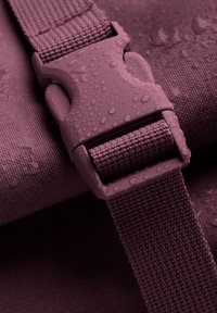 Detail fialového nepromokavého vaku s texturovaným popruhem a plastovou sponou, na kterém jsou kapičky vody, což zdůrazňuje jeho odolný materiál a design.