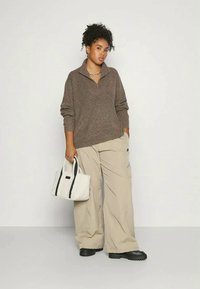 Woman with curly hair in brown zip-up sweater, beige wide-leg pants, black shoes, holding white tote bag, standing against plain light background.