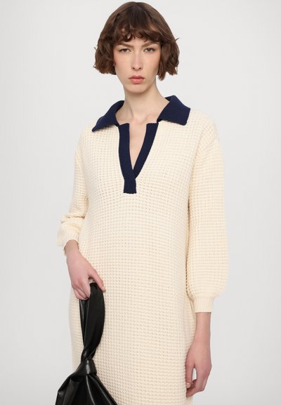 Rochie tricotată în alb murdar cu guler bleumarin, având decolteu în V și mâneci lungi, voluminoase. Modelul ține o geantă neagră.