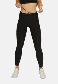 Schwarze, hoch taillierte Leggings aus dehnbarem Material mit seitlichen Taschen und einem Zugband am Bund. Getragen mit weißen Turnschuhen.