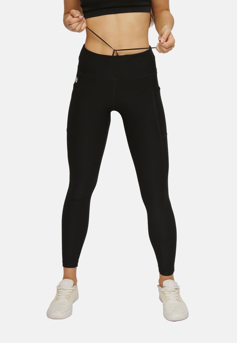 Schwarze, hoch taillierte Leggings aus dehnbarem Material mit seitlichen Taschen und einem Zugband am Bund. Getragen mit weißen Turnschuhen.