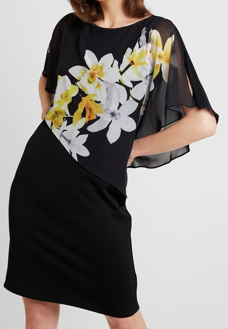 Robe noire avec un voile transparent à imprimé floral blanc et jaune. Le modèle comprend des manches courtes et fluides ainsi qu'une jupe noire cintrée.