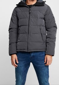 Graue Steppjacke mit durchgehendem Reißverschluss, gepolstertem Design und verstellbarer Kapuze. Verfügt über zwei seitliche Taschen und elastische Bündchen für Wärme.