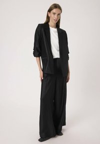 Schwarzer Strukturanzug mit lockerer Jacke und weit geschnittenen Hosen. Darunter ein weißes T-Shirt, minimalistisches Design, glattes Material und dezenten Glanz.