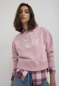 Felpa rosa chiaro con la scritta "Rosé Lovers" in font metallico argento, abbinata a una camicia di flanella a quadri rosa e bianca sotto.