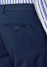 Pantaloni di cotone blu navy con una tasca posteriore caratterizzata da un bottone nero e un piccolo etichetta con la scritta "HACKETT". Texture liscia, design su misura.
