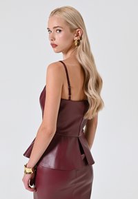 Burgunderfarbene Peplum-Bluse aus Kunstleder, mit dünnen Trägern und tailliertem Schnitt. Glatte Textur und subtiler Glanz mit einem Reißverschluss auf der Rückseite.