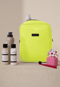 Neon gele gestreepte toilettas met zwart logo, drie flessen shampoo en conditioner, roze container met bloem, en een rode tandenborstel.