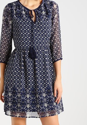 Dunkelblaues Kleid mit floralem Muster, dreiviertel Ärmeln, gebundenem Ausschnitt und strukturiertem unteren Saum. Verfügt über einen Quastenakzent.
