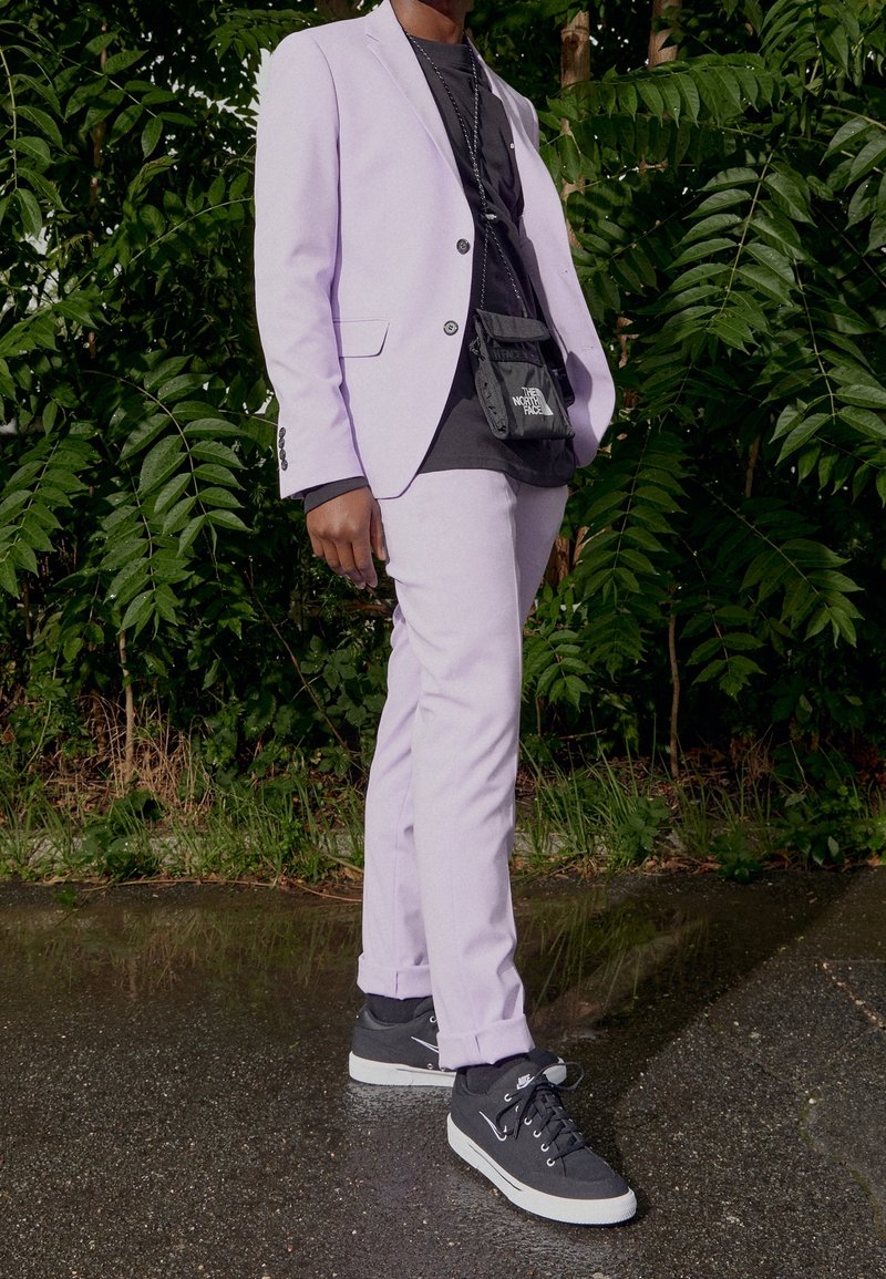 Lila kostym med en figursydd kavaj och smala byxor, bärs över en svart t-shirt. Matcher med svarta sneakers med vita sulor.