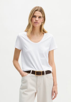 Blondynka ubrana w białą koszulkę z krótkim rękawem wpuszczoną w jasnobeżowe spodnie z ciemnobrązowym paskiem, stoi z jedną ręką w kieszeni.