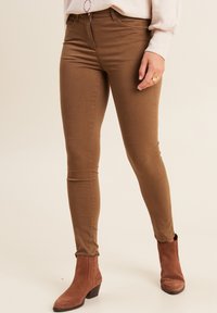 Pantaloni marroni slim-fit in tessuto liscio, con chiusura a zip standard, tasche frontali e orlo leggermente sfilacciato. Indossati con stivali alla caviglia marroni.