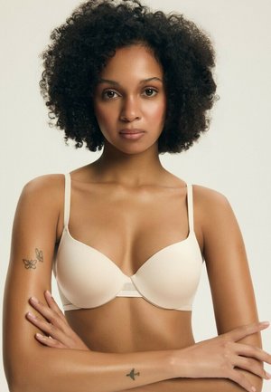 Kobieta z kręconymi włosami, ubraną w beżowy biustonosz, z założonymi skrzyżowanymi ramionami, prezentująca tatuaże motyla i samolotu na górnej i dolnej części ramion.