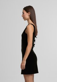 Černé sametové šaty s přiléhavým střihem, bez rukávů a ozdobnými stužkami podél zad. Hladká textura.