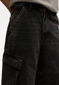 Zwarte denim cargo broek met een relaxed fit, voorzien van zijzakken en een subtiele textuur. G gedragen met een lichtgrijs shirt.