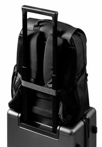 Black backpack with a padded back resting on a black wheeled suitcase. Retractable handle extends from the suitcase for easy transport.