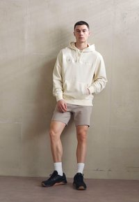 Krémová mikina s kapucňou a predným vreckom, doplnená bledými šortkami. Model nosí čierne topánky a biele ponožky.