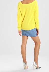 Helles gelbes Strickoberteil mit weitem Ausschnitt, kombiniert mit einem blauen Jeans-Minirock. Das Model trägt silberne High Heels.
