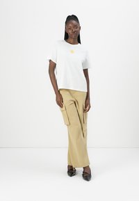 Kvinne som står iført hvit t-skjorte med gul solgrafikk, beige cargo-bukser og svarte spissede sko mot hvit bakgrunn.