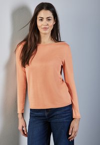 Długi rękaw, koralowy sweter z dekoltem w łódkę i ściągaczami, zestawiony z ciemnoniebieskimi dżinsami. Gładka faktura, dopasowany krój.