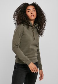 Felpa verde oliva realizzata in morbido tessuto, con cappuccio regolabile, polsini a coste e una vestibilità comoda. Indossata con jeans neri.
