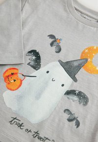 T-shirt gris à manches longues avec un fantôme portant un chapeau de sorcière, tenant une citrouille, entouré de chauves-souris et d'un motif de croissant de lune.