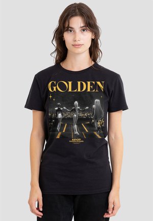 Woman wearing black T-shirt with gold text "GOLDEN" and graphic of three stylized female figures walking on stage toward a crowd.