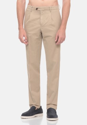 Uomo che indossa pantaloni beige a gamba dritta e scarpe nere slip-on in piedi su uno sfondo semplice.