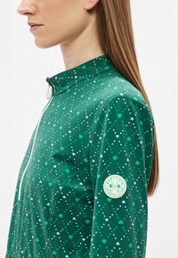 Πράσινο μπουφάν με φερμουάρ, διακοσμημένο με μοτίβο καρδιάς και κουκκίδων, φτιαγμένο από λεία ύφασμα, με ετικέτα λογότυπου στο μανίκι.