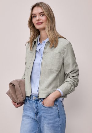 Jeune femme aux cheveux blonds portant une veste à fermeture éclair claire à motifs, une chemise bleu clair et un jean bleu, tenant un manteau beige plié dans une main.