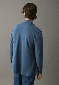 Uomo in piedi di spalle, vestito con una camicia a maniche lunghe blu e jeans blu, su uno sfondo grigio uniforme.