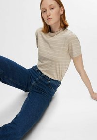 Camisa de rayas beige de manga corta, con escote redondo, combinada con vaqueros de tiro alto de color azul oscuro, sentada sobre un fondo claro.