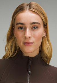 Brown zip-up athletic top with fitted design, textured fabric, and visible stitching. Model has medium-length blonde hair and simple earrings.